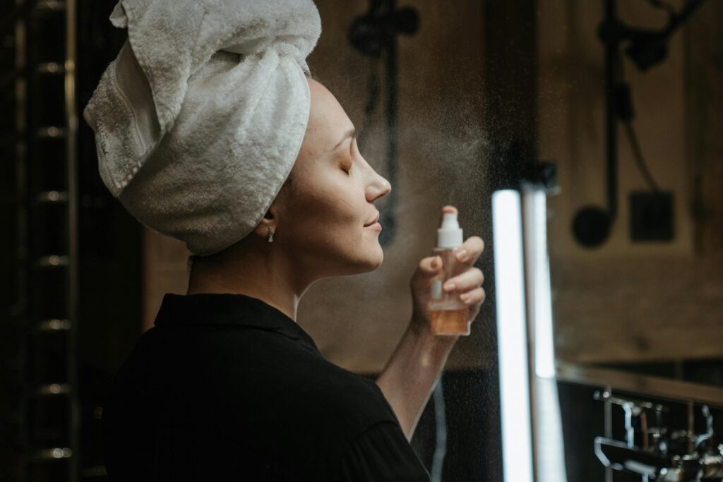 Adult woman with towel on head applies a refreshing facial mist in her skincare routine.